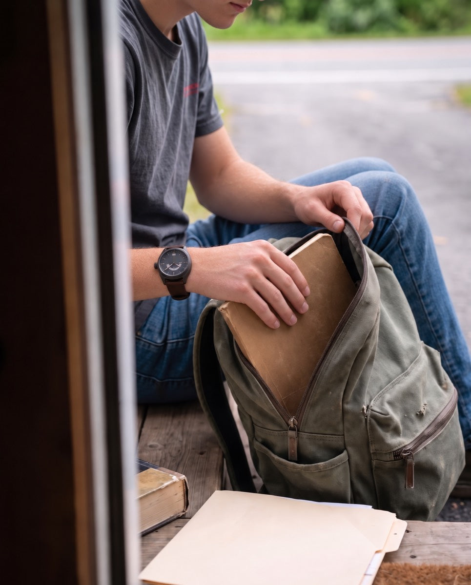 Close on his hands packing a notebook into a canvas backpack on the front step, the watch on his left wrist.
