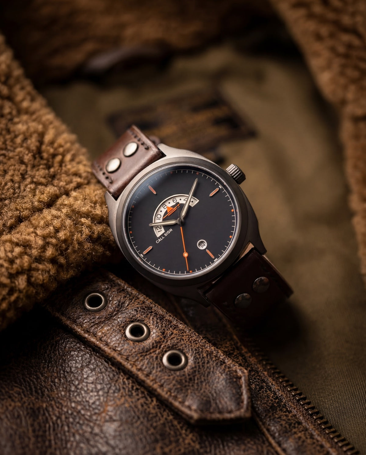A Discovery with a dark dial and the orange crescent GMT window at 12, on a brown riveted leather strap, resting on the shearling collar of a leather flight jacket.
