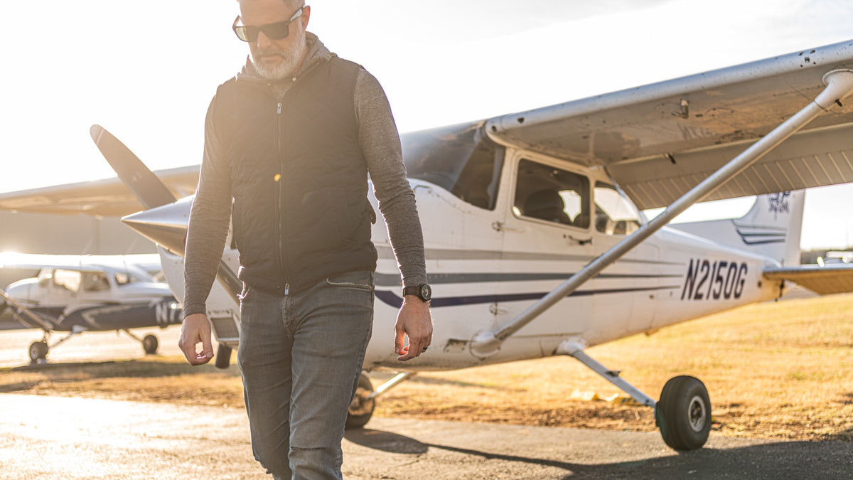 A pilot in a quilted vest walks across the ramp toward the camera in low evening light, two Cessnas behind him, a Discovery on his left wrist.
