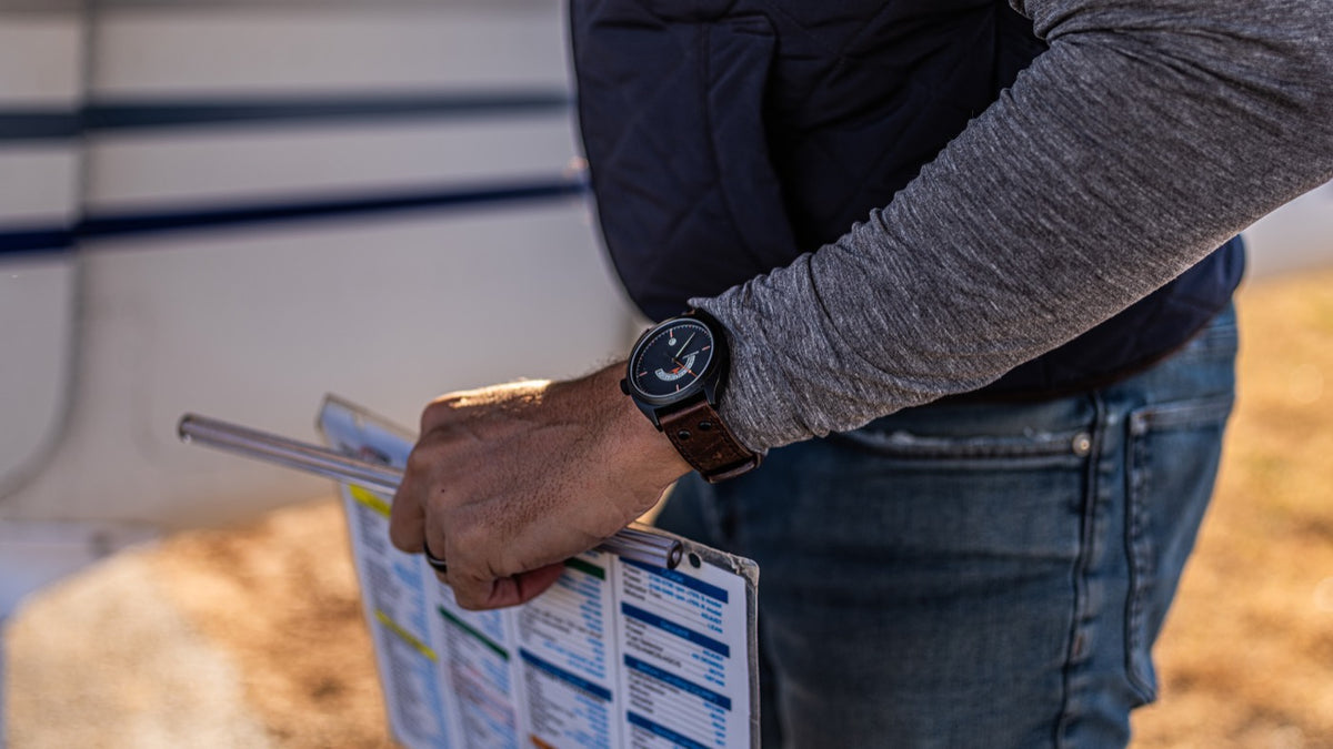 A pilot's hand holds a checklist against the wing strut of a Cessna during preflight, the Discovery on his wrist.