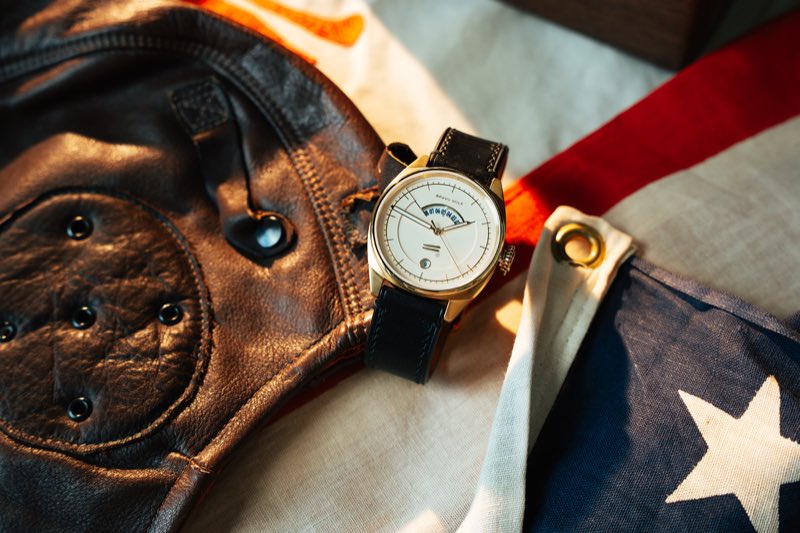 The Bishop, cream dial in a gold-tone case on black leather, resting on a leather flying helmet and an American flag.